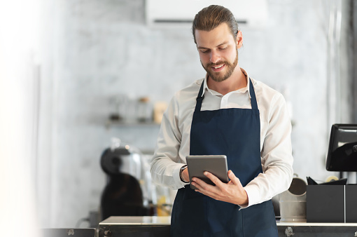 Portrait of a Hipster barista in blue apron using digital tablet standing at the bar of the modern cafes. Smiling attractive Bearded man receiving hot drink online orders with service mind