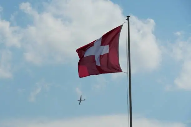 Flapping flag of Switzerland displays a white cross in the centre of a square red field. Flag is attached to flagpole and in background is visible gliding sailplane in blue sky with white clouds.