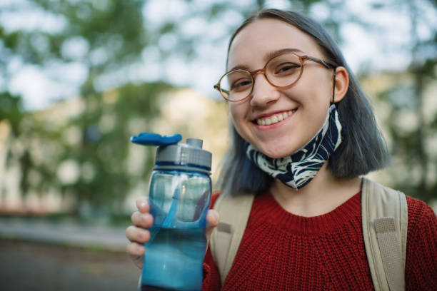 Teenage girl in reusable protective face mask drinking water outdoors in campus or schoolyard. New normal and Back to school concept after COVID-19 quarantine Female high school or college student outdoors during COVID-19 pandemic. blue reusable water bottle stock pictures, royalty-free photos & images