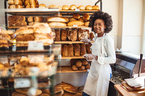 She will have you coming back for more. Gorgeous young woman in her bakery smiling cheerfully to the camera. Smiling young woman selling bread in the bakery. Happy african baker woman