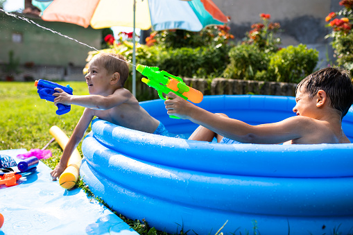 Two small boys are shooting someone with water guns from a paddling pool