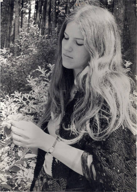 Pensive young woman in headphones sitting under tree with thick trunk, listening to music and making notes in diary while looking aside