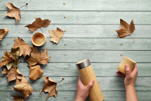 istock Zero waste eco friendly insulated bamboo flask with bamboo cups. Trendy flat lay with hands holding the flask and natural bamboo cup on aged blue mint wooden table with Autumn sycamore leaves. 1269522952