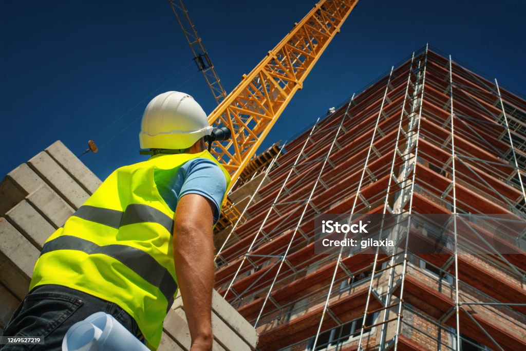 Construction engineer supervising building process Closeup construction engineer standing in front of a building. He's doing some calculation and inspection by looking at construction plan and using binoculars. Construction Industry Stock Photo