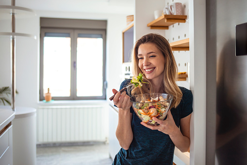 Photo of cheerful woman standing with salad dish at home kitchen. Beautiful girl eating fresh salad at modern apartment. Closeup young woman enjoying vegetarian food indoors.