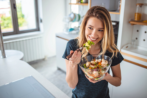 Starting day with healthy food. Beautiful young woman eating salad and looking away while standing in kitchen at home