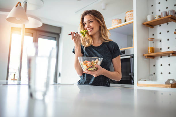 What I eat is who I am Photo of young woman enjoying a delicious salad while standing in her kitchen at home during the day. healthy eating stock pictures, royalty-free photos & images