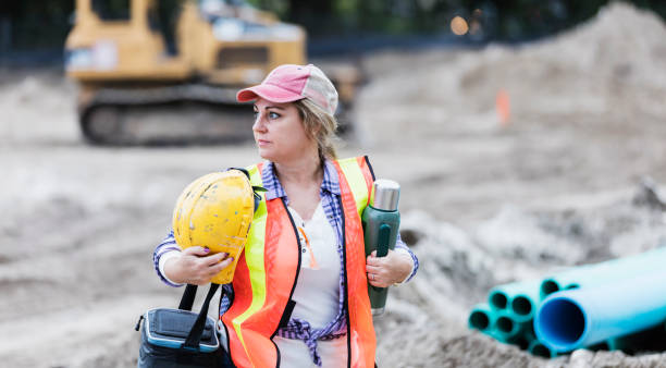 Mature woman working at construction site, on break A mature Hispanic woman in her 40s carrying a hard hat and wearing a reflective vest, working at a construction site. An earth mover, large tubes and other construction material are out of focus in the background. She is on a break, carrying an insulated drink container and lunch box. construction lunch break stock pictures, royalty-free photos & images