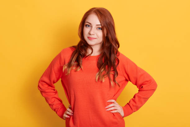 istock Beautiful woman with red wavy hair, looking directly at camera while keeping hands on hips, posing in orange casual sweater against yellow background. 1269546206