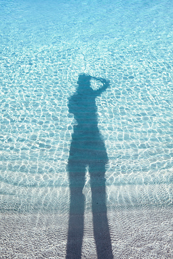 Young female taking an auto photo reflected on the water on the shore of a beach in Spain. Shadow of a person’s silhouette