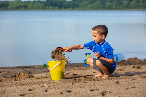 Close-up of a young four year old boy tossing sand from his shovel into a yellow bucket on the beach. Taken on a summer morning at the lake.