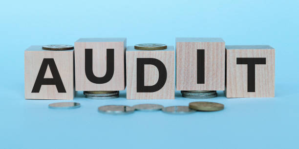 istock AUDIT word written on wooden blocks on stacks of coins on light blue backround. Selective focus. Top view. 1269552004