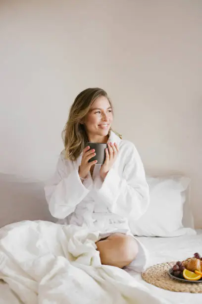 Photo of Young beautiful woman wearing white bathrobe having breakfast in bed with coffee and croissant and fresh fruits in cozy bedroom.