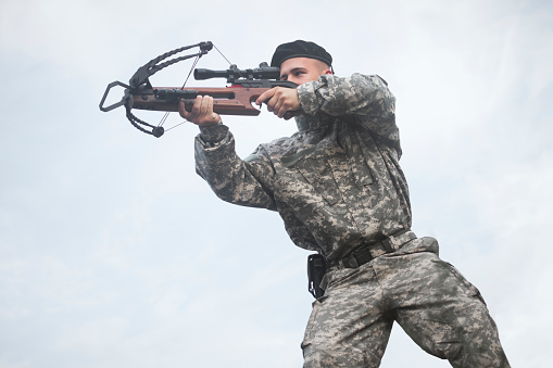 The army soldier in military uniform is aiming and shooting a crossbow weapon on cloudy white sky background outdoors.
