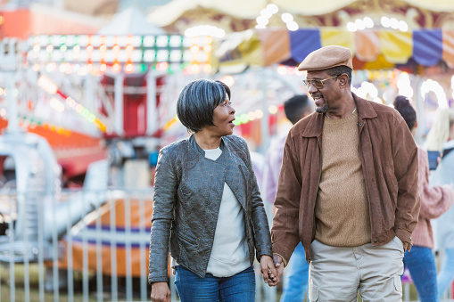 A senior African-American couple having fun together at a traveling carnival. They are looking at each other, holding hands and conversing as they walk. They are both in their 60s.