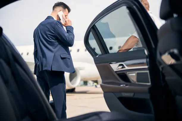 Young businessman talking on the phone standing by the taxi with an open door getting picked up from the flight