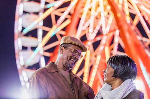 A senior African-American couple in their 60s having fun together at a traveling carnival. An amusement park ride is out of focus in the background.