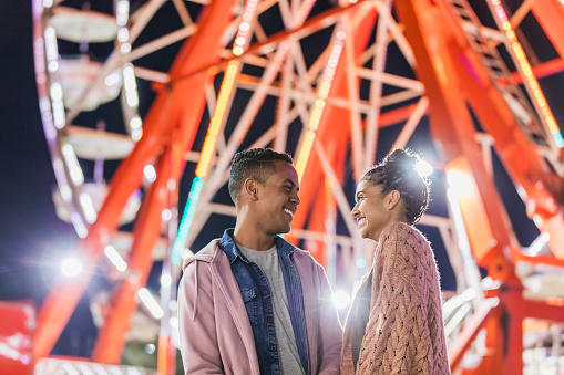A young couple in their 20s having fun together at a traveling carnival. An amusement park ride is out of focus in the background. He is African-American and his girlfriend is mixed race African-American and Caucasian.