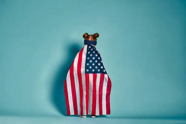 Photo of American flag. Back view little patriotic happy girl wrapping in usa national flag on blue background. National 4 july. Memorial day.