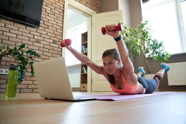 Active fit sporty young woman holding dumbbells doing superman fitness exercise training abs and back at home on mat watching online video training tutorial sport live stream workout class on laptop.