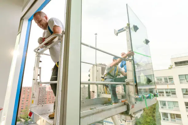 Team of blue-collar workers replacing a broken window in the office building. High-altitude work on the lifting platform which is placed outside.