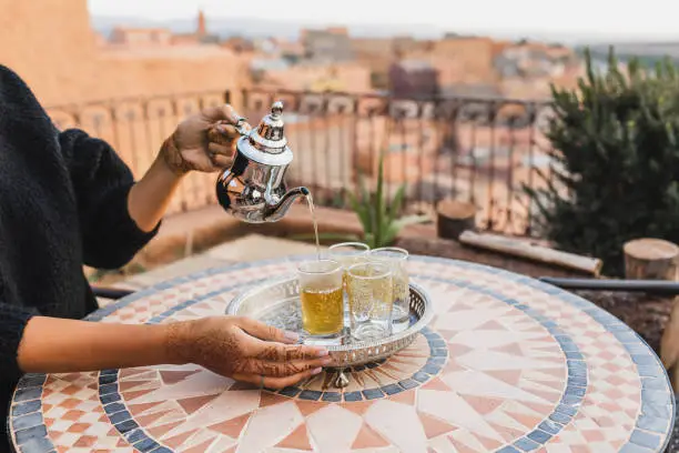 Photo of Woman hand pouring traditional moroccan mint tea in glasses. Vintage silver tray and teapot. Round mosaic table. Morocco hospitality.