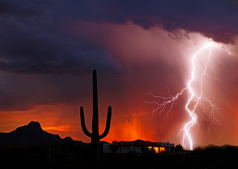 Powerful lightning strike near a house, saguaro cactus, and mountain backlit by an orange sunset and falling rain with interior lights visible in the house
