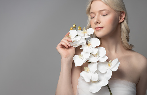 Charming blonde lady with orchid closing eyes and touching petals while standing against gray background