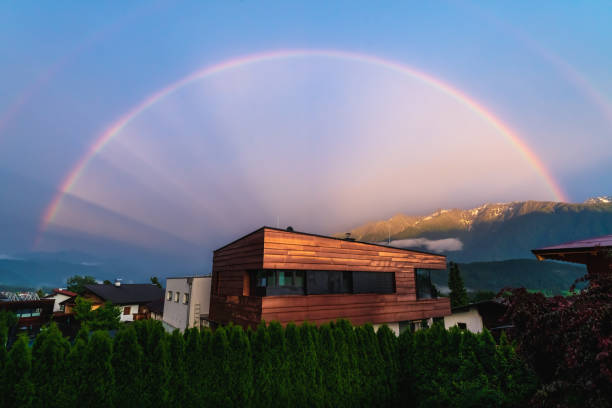 Wildermieming, Mieminger Plateau, Tyrol, Austria - 27 June 2020: Scenic full Rainbow over modern copper building in Austrian alp mountains village during sunset