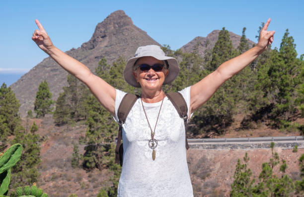 istock Happy active senior woman with hat and sunglasses enjoying excursion with backpack in mountain landscape in Tenerife  - concept of summer vacation for an elderly people 1269649017