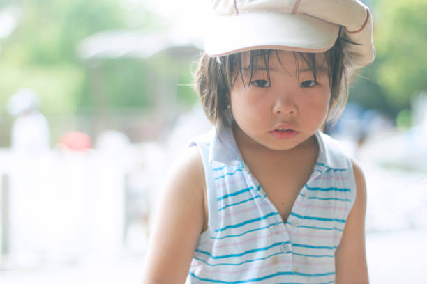 A baby girl is standing outdoors. She wears casual clothing and hat. She is looking at here.