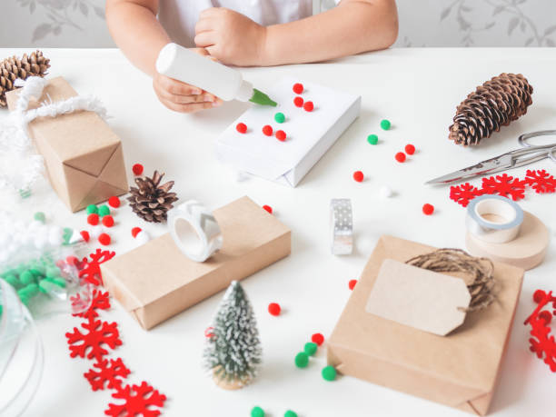 Kid wraps handmade Christmas presents in craft paper with colorful pompons and snowflake ribbons. Child prepares gifts for New Year celebration. Peaceful leisure activity before winter holidays. los niños envuelven regalos de navidad hechos a mano en papel artesanal con pompones coloridos y cintas de copo de nieve. el niño prepara regalos para la celebración del año nuevo. actividad de ocio tranquilo antes de las vacaciones de invierno.
