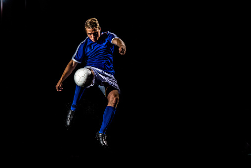 Caucasian male soccer player in a blue and white kit about to kick the ball in mid-air on black background.
