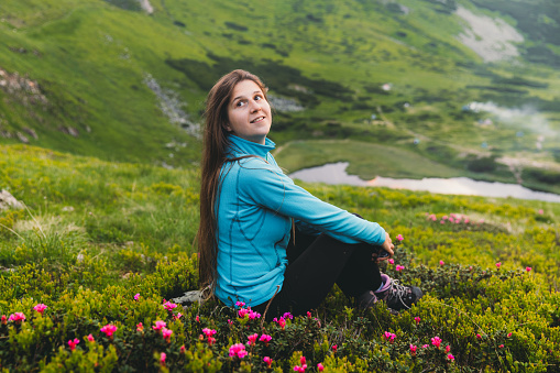 Happy young woman traveler resting after the hiking trip at Carpathian Mountains sitting at the beautiful green meadow with the bright oink flowers in the wilderness area