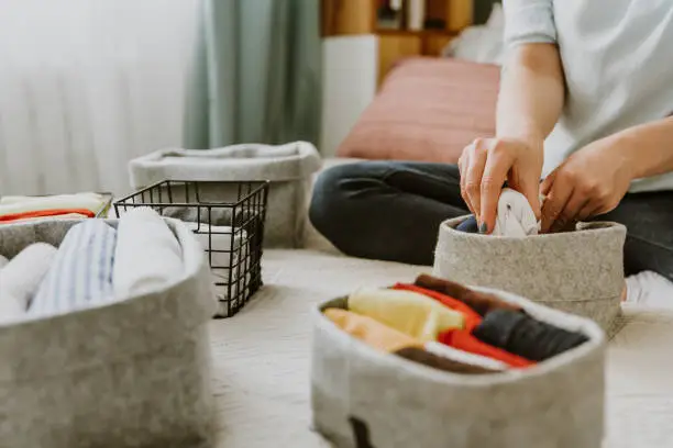 Photo of Woman organizing clothes in wardrobe, putting shirts in boxes, baskets into shelves. Clothes neatly folded after laundry. Concept of minimalist lifestyle