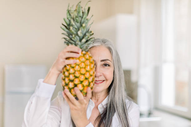 middle age beautiful woman wearing white shirt, holding pineappe and hiding half of her face, looking at camera with happy smile while standing in modern kitchen at home. healthy food - half smile imagens e fotografias de stock