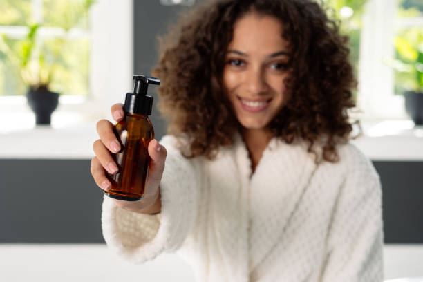 Afro american woman in bathrobe spending morning in bathroom Beauty and wellness concept. Selective focus on bottle with dispenser in afro american woman hands. Happy african girl standing in bathrobe at bathroom, holding natural skincare or body care cosmetics shower gel photos stock pictures, royalty-free photos & images