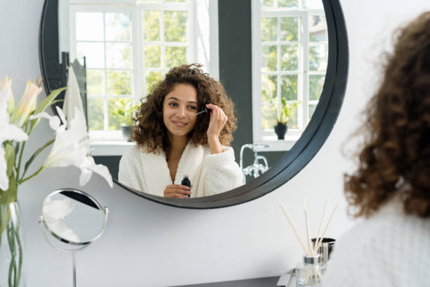 Young adult african american woman in bathrobe at bathroom Concept of beauty procedure at home. Happy young adult african american woman holding pipette with anti wrinkle serum near eye, looking at mirror wearing in bathrobe, sitting at bathroom serum sample stock pictures, royalty-free photos & images