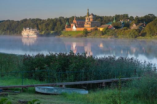 A tourist motor ship against the background of the male Nikolsky monastery in Staraya Ladoga in the Leningrad Region on a foggy morning on the Volkhov River. Traveling on the rivers of Russia