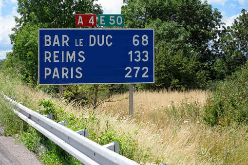 A road sign to the towns of Bar le Duc, Reims and Paris, 272 km away, on an abandoned highway among green trees and grass on the wayside. Sunny day. Copy space.