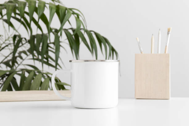 istock Enamel mug mockup with workspace accessories on a white table and a palm plant. 1269709177