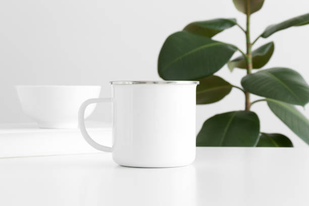 istock Enamel mug mockup with workspace accessories on a white table and a ficus plant. 1269710235