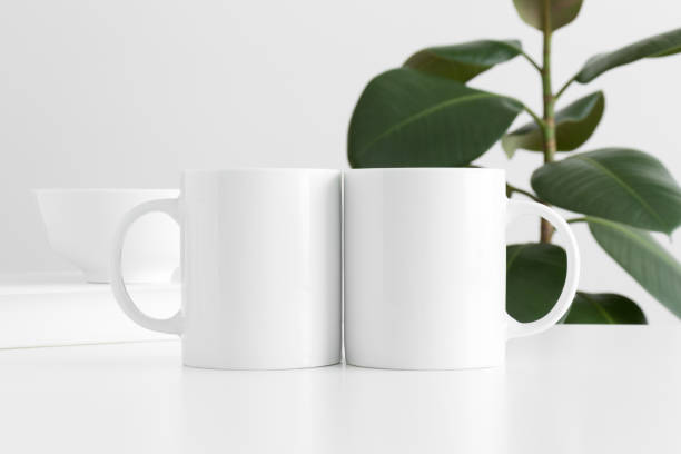 istock Two white mugs mockup with workspace accessories on a white table and a ficus plant. 1269710243