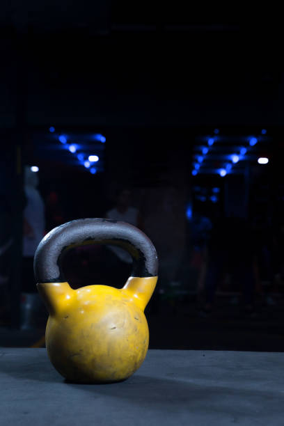 gelbe kettlebell mit schwarzem griff auf schwarzem boden mit fitness-studio hintergrund und bokeh lichter low key dunkle beleuchtung mit kopierraum - gym yellow muscular build dumbbell stock-fotos und bilder