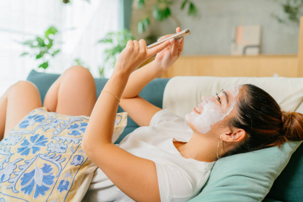 женщина носить маску красоты лица лежа на диване и наслаждаясь использованием своего смартфона в гостиной у себя дома - asian spa treatment стоковые фото и изображения