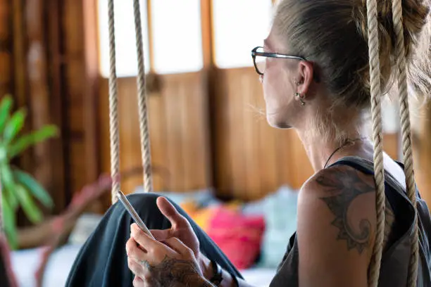 One mid adult Woman is sitting on an indoor swing holding her smart phone.  She is day dreaming and looking away from the phone.