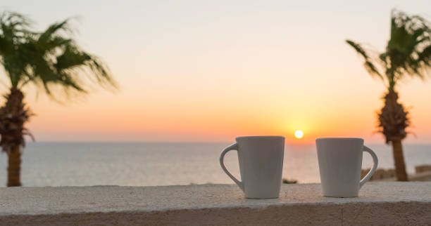 2 white cups against the background of the rising sun, the sea and two palms with an empty spot on the left side stock photo
