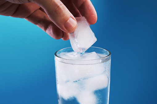 person putting ice to water glass close up view over blue background. copy space. refreshment beverage. healthy lifestyle concept.