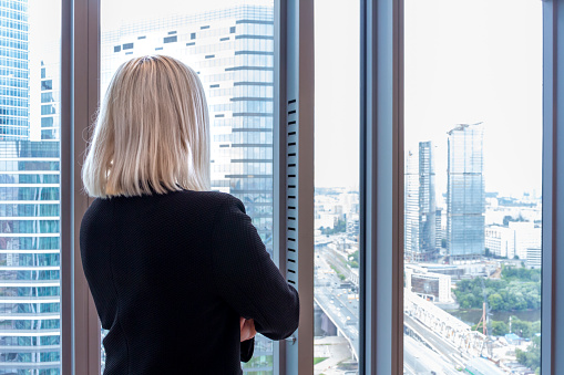 Сaucasian blonde hair woman stands in office near the window. Blurred office buildings are visible through window. Back view. Copy space for your text. Theme of Successful women in business.