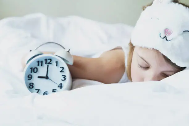 Photo of Bedtime. Little girl goes to sleep early evening with alarm clock on 9 p.m., wearing white cat, bunny sleep mask. Soft bed with white linen.Child sleep regime hours,daily routine.Sweet dreams concept